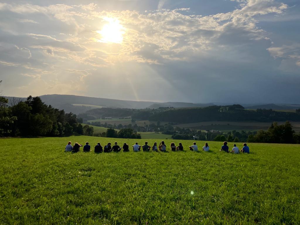 Řada dětí sedí na louce a dívá se do krajiny